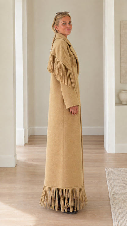 Side profile view of woman wearing long camel overcoat with shoulder and hem fringe