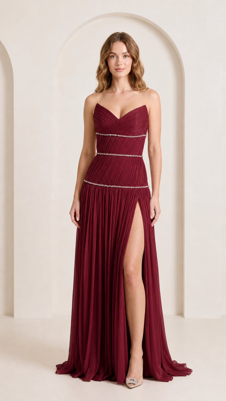 Front view of woman wearing rich burgundy strapless pleated maxi gown with rhinestone waist trim